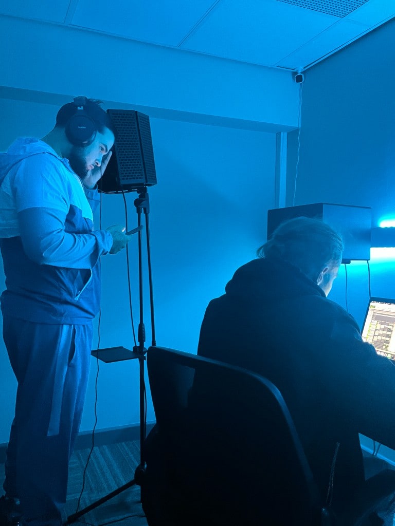Artist recording vocals at Warrington Music Studio with engineer at the desk, pop filter and acoustic shield in blue session lighting.